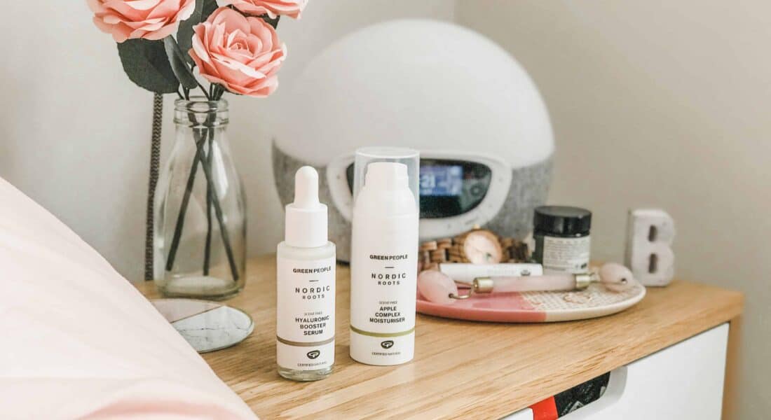 Green People Nordic Roots Hyaluronic Booster Serum and Apple Complex Moisturiser on a bed side table
