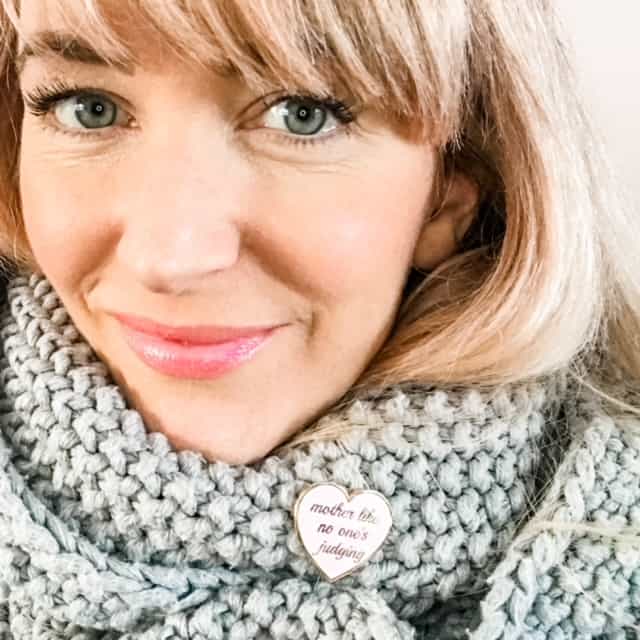 Becky Pink wearing a grey scarf with a pale pink and gold heart-shaped enamel badge with the words "Mother like no one's judging". She's smiling and looking directly at the camera 