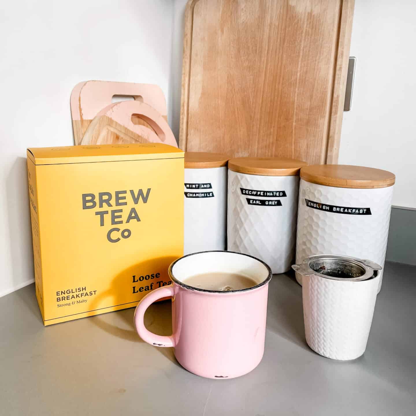 A box of Brew Tea plastic-free tea leaves and a cup of tea made with it, in front of some canisters of tea on a worktop