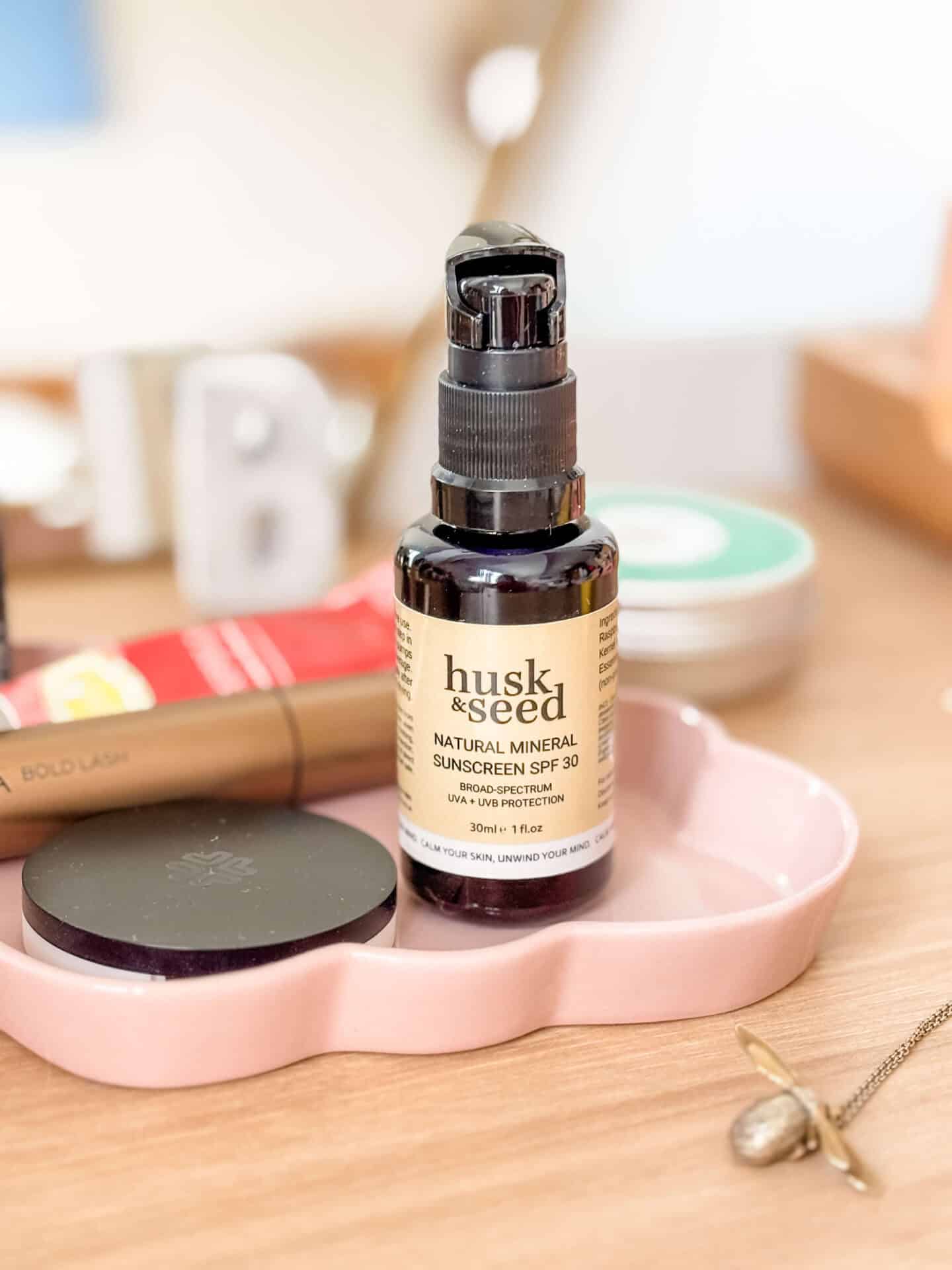 A glass bottle of Husk & Seed Natural Mineral Sunscreen SPF30 on a pink plate surrounded by clean make-up