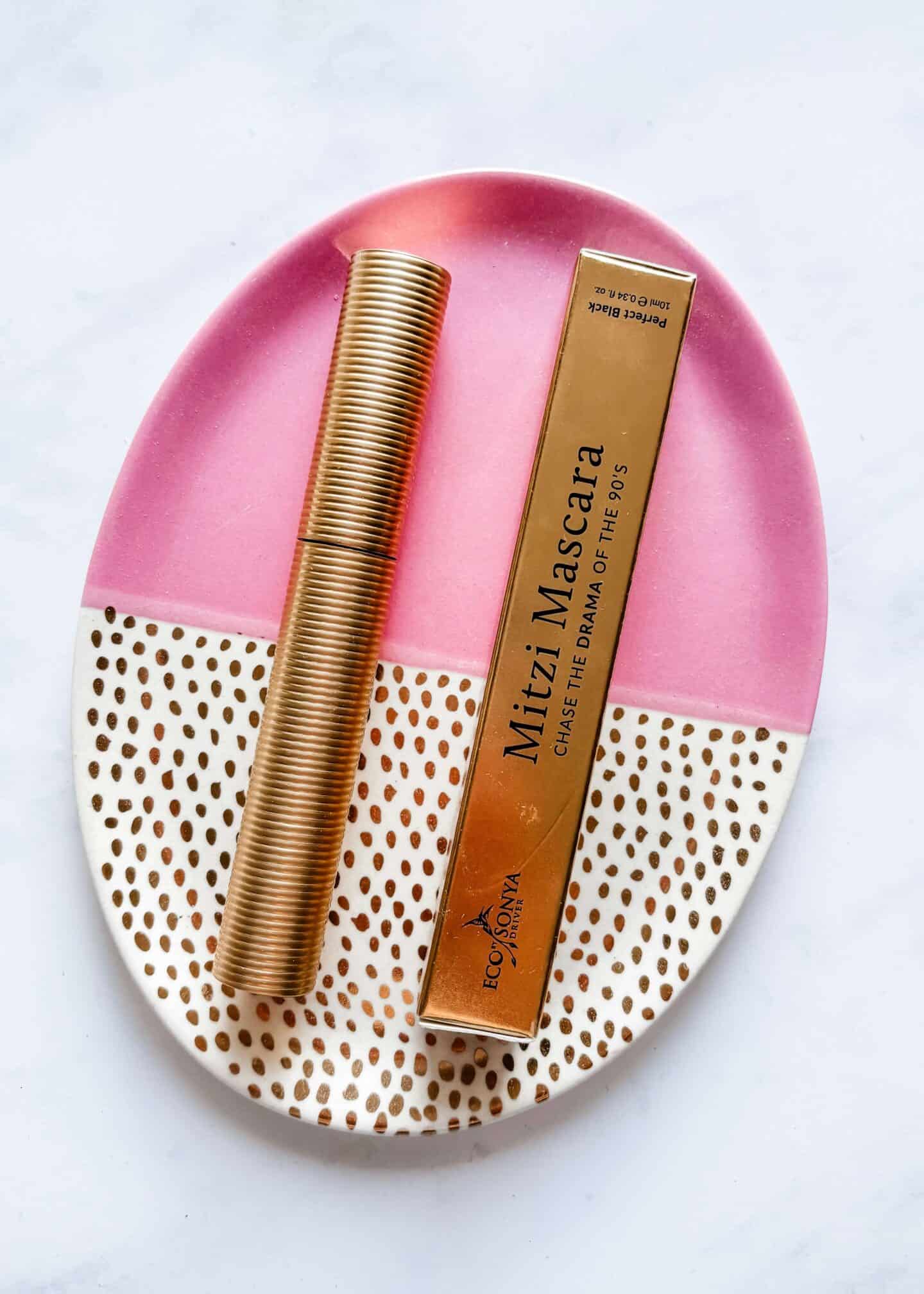 A tube of Eco by Sonya Driver Mitzi Mascara and it's golden box on a pink and gold plate