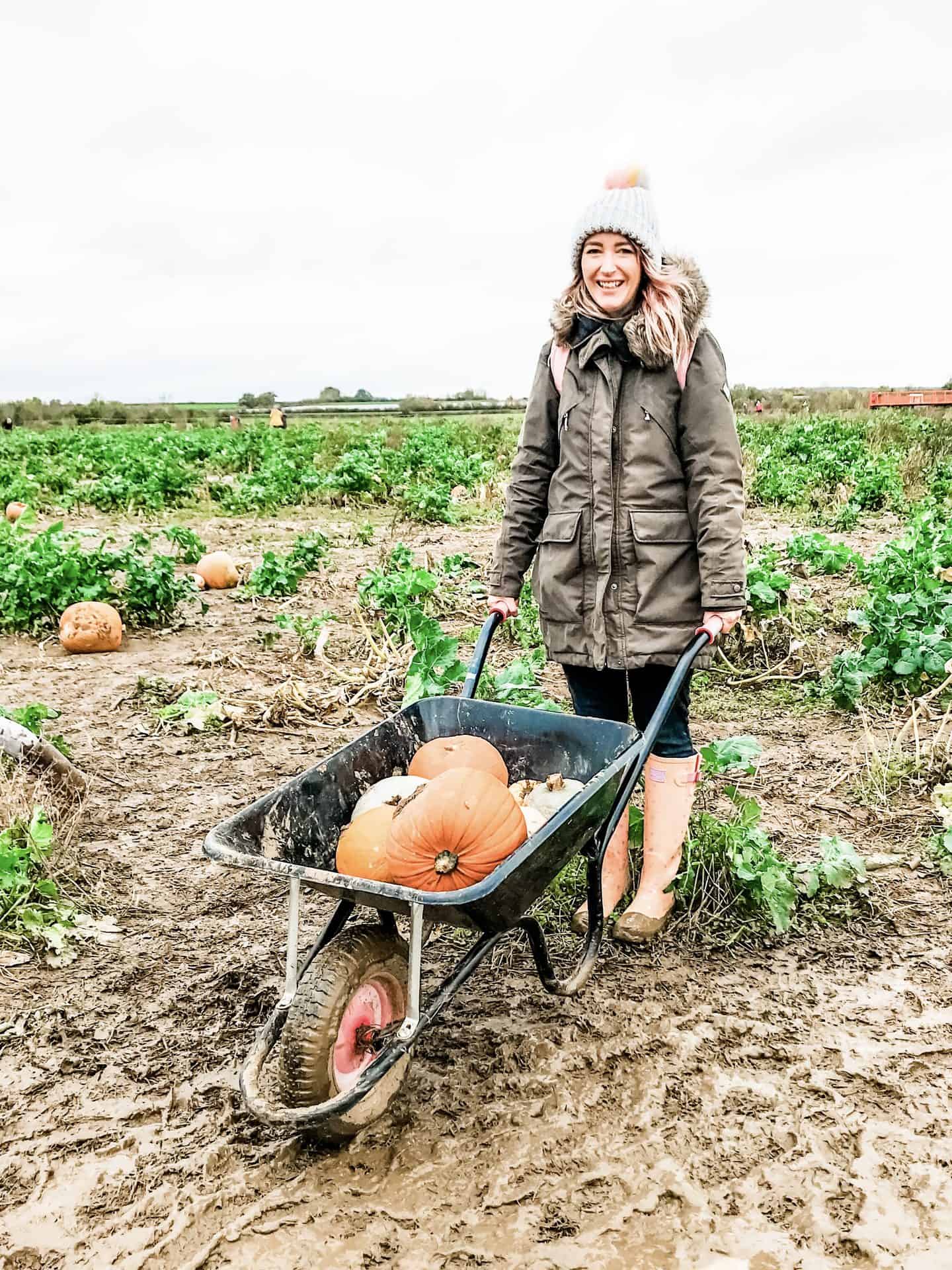 A white woman standing in a muddy field with a wheelbarrow containing freshly-picked pumpkins, laughing as she triesto create a lower-waste Halloween